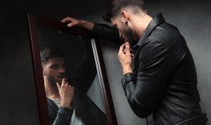 Seorang pria melihat ke cermin, dengan bayangan yang tampak lebih besar dan lebih kuat dari aslinya, menggambarkan konsep narsisme dan distorsi persepsi diri.